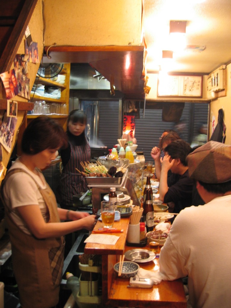 A small, crowded local izakaya in Tokyo with a chef cooking behind a narrow wooden counter