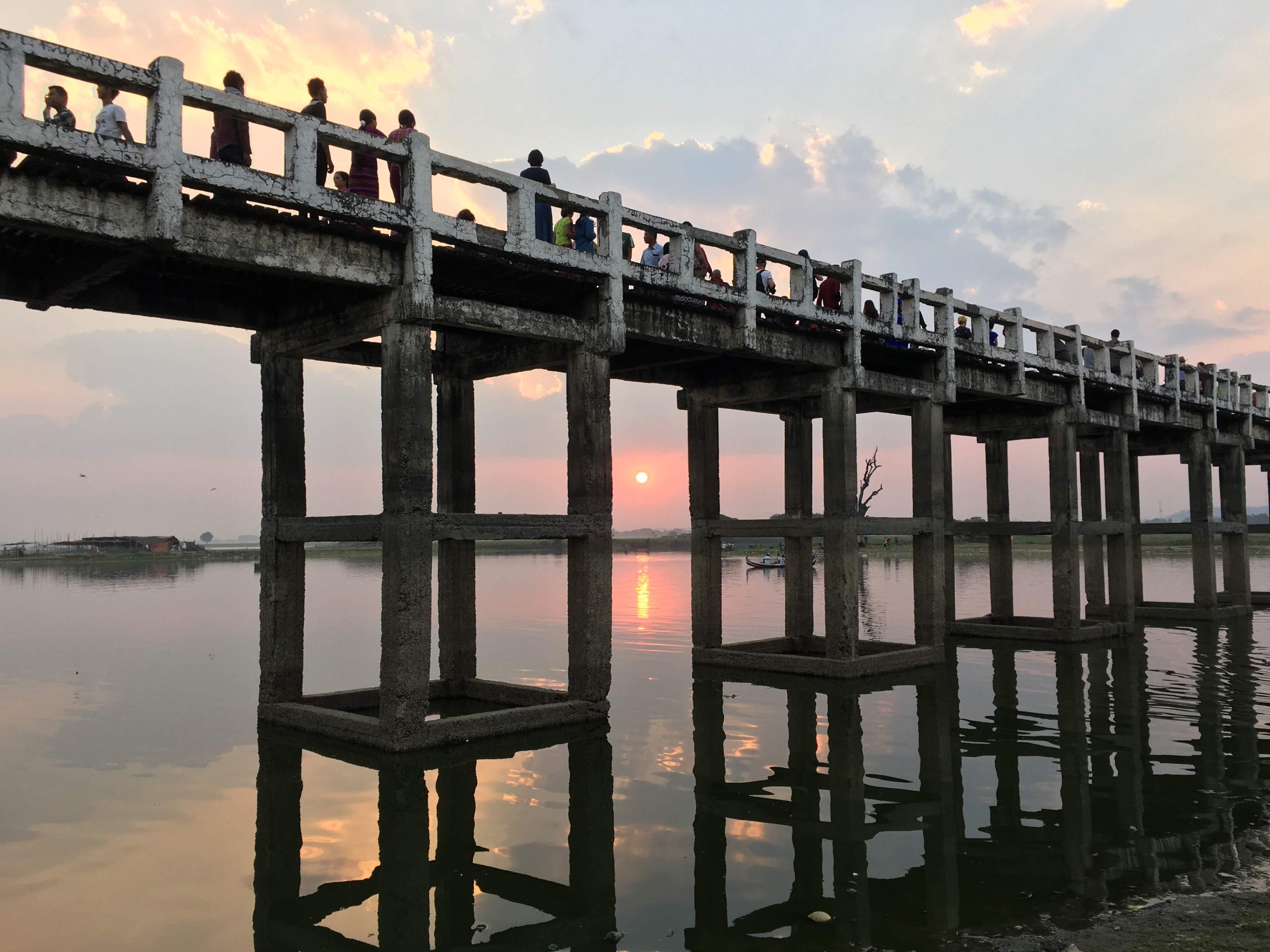 A stunning sunset over Taungthaman Lake, featuring the dark silhouettes of people walking across the wooden U Bein Bridge against a vibrant orange and red sky.